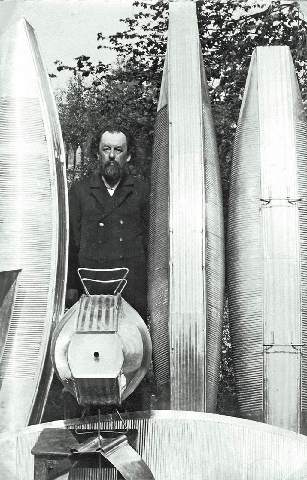 Konstantin Tsiolkovsky, bearded and in a dark coat, standing among tall corrugated-metal airship models in a garden