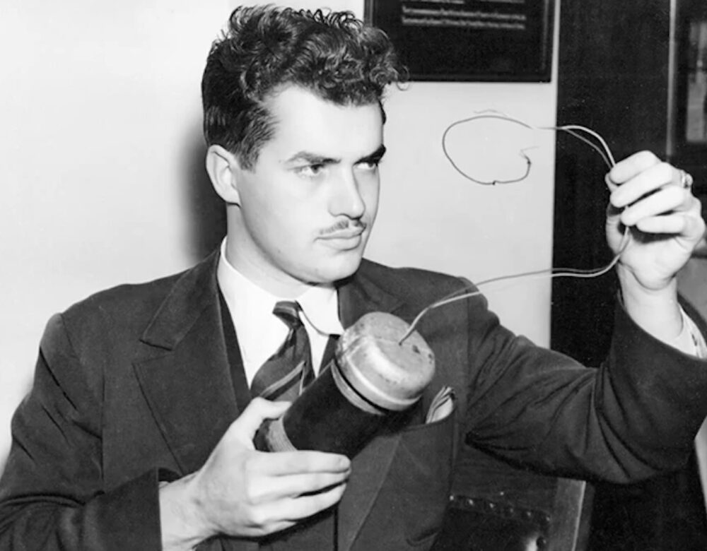 A young Jack Parsons in a suit holding up a cylindrical bomb casing with wires trailing from it
